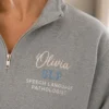 Close-up of a grey quarter-zip with Olivia in blush script, SLP in blue and SPEECH LANGUAGE PATHOLOGIST