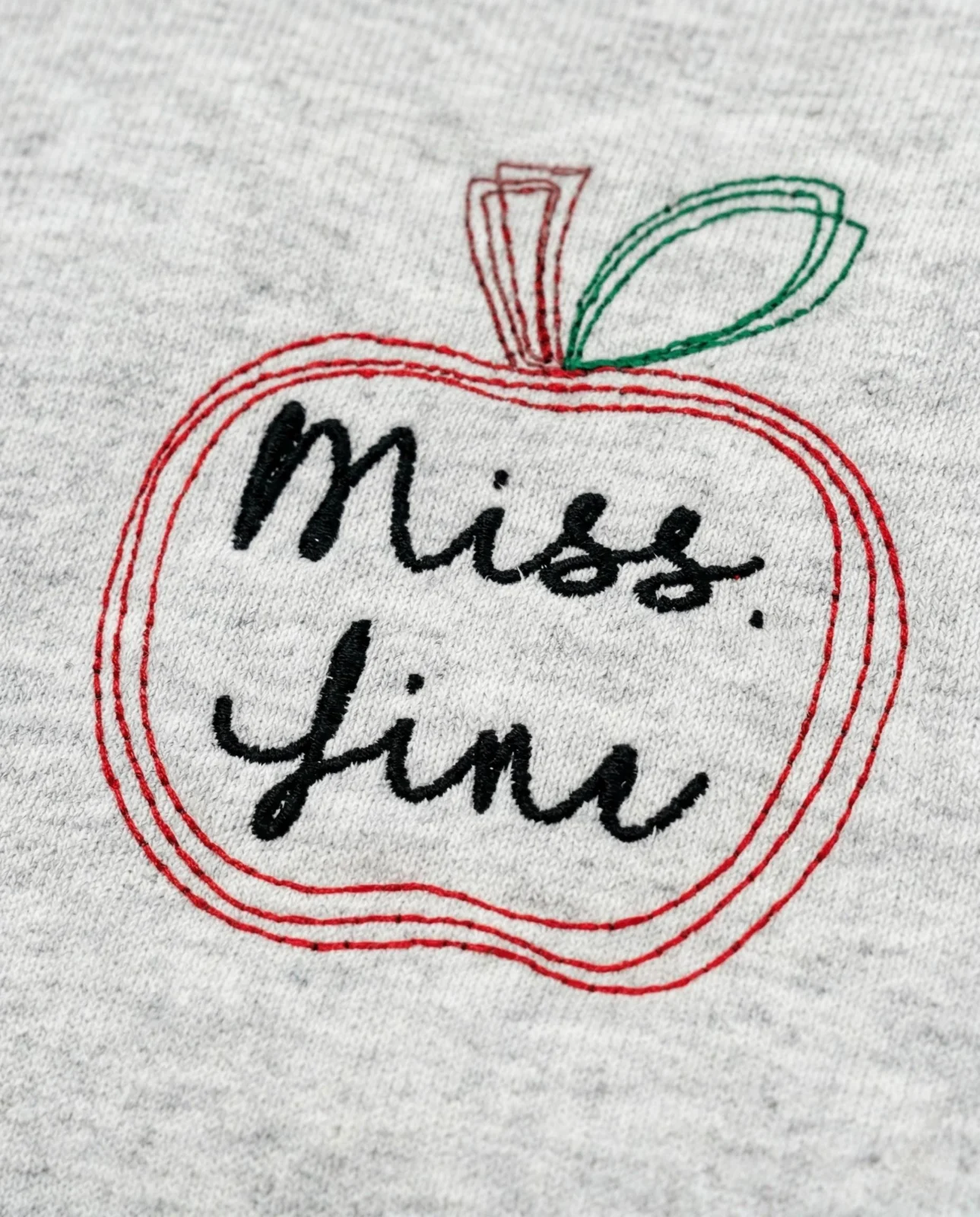 Close-up of a red outline apple with a green leaf framing the name Miss. Jini in black script embroidery