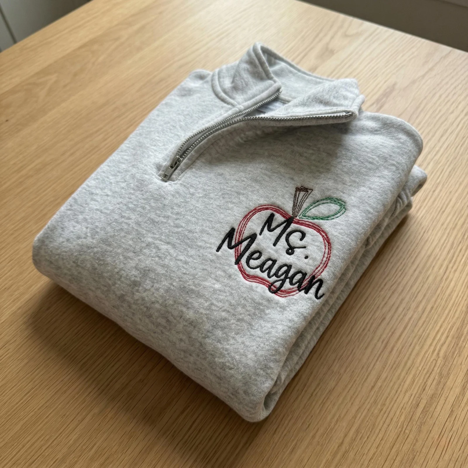 Folded ash quarter-zip sweatshirt with a red sketched apple outline and Ms. Meagan in black script on the left chest