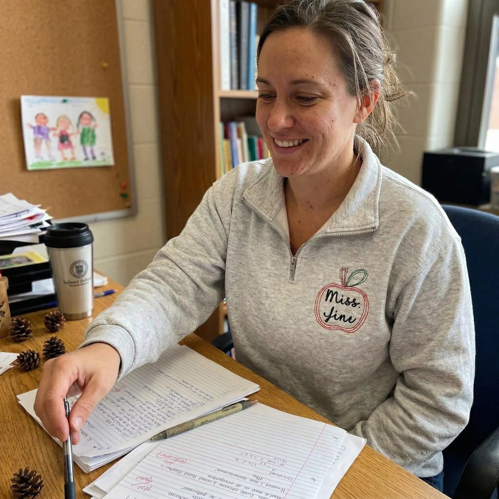 Smiling teacher at her desk wearing an ash quarter-zip embroidered with an apple outline and Miss. Jini