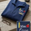 Folded navy quarter-zip with a ruler, red apple, Miss. Lizze in white and a pencil embroidered on the left chest