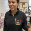 Smiling teacher in a classroom wearing a black quarter-zip embroidered with Ms. Jane, a ruler, apple and pencil