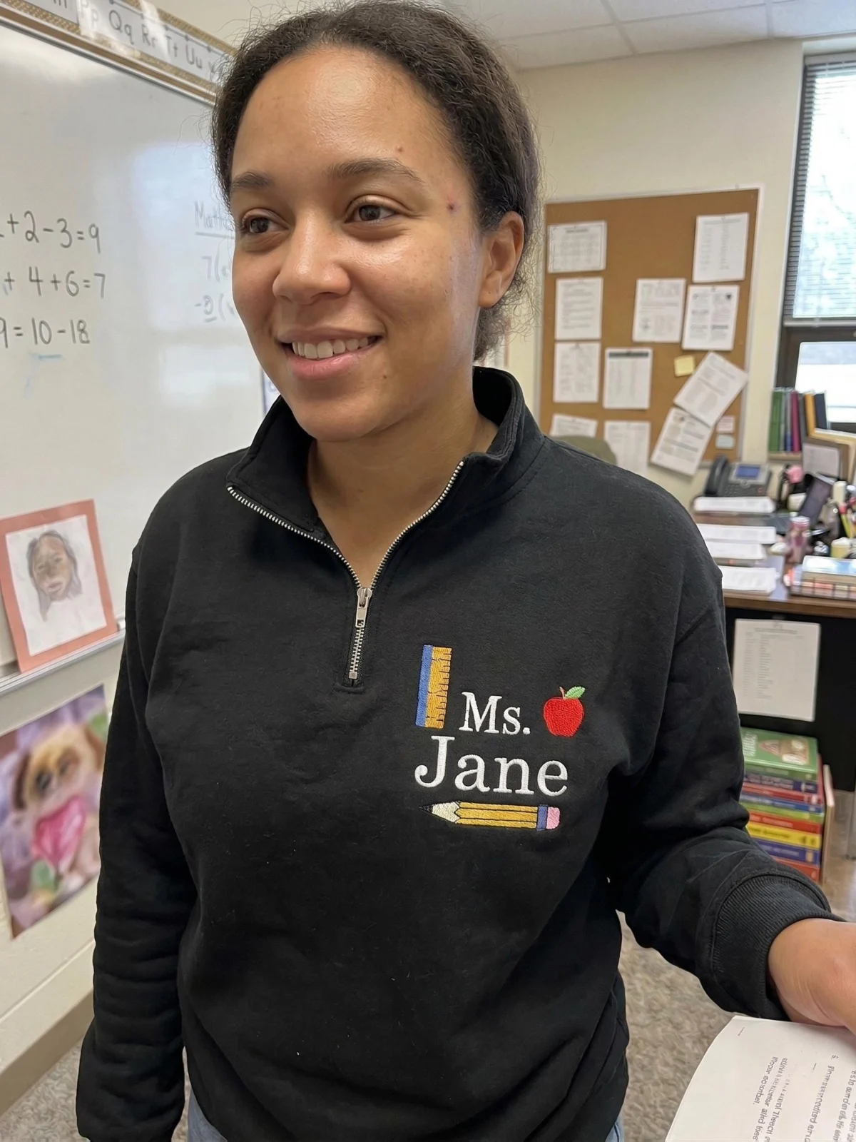 Smiling teacher in a classroom wearing a black quarter-zip embroidered with Ms. Jane, a ruler, apple and pencil