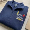 Navy quarter-zip sweatshirt folded on a desk with a teacher name badge of ruler, apple, script name and pencil