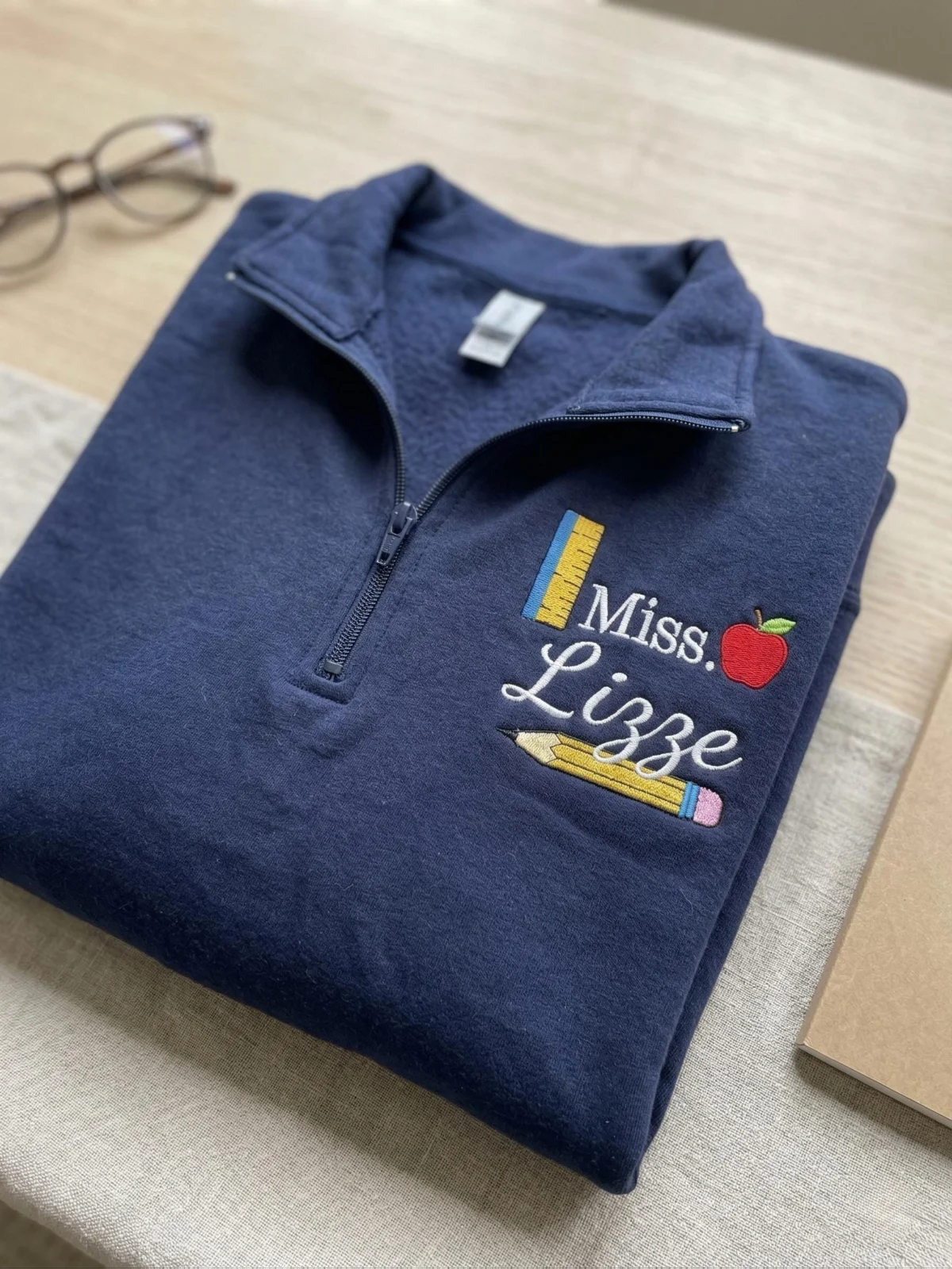 Navy quarter-zip sweatshirt folded on a desk with a teacher name badge of ruler, apple, script name and pencil