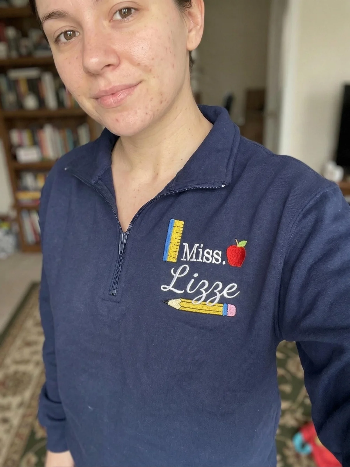 Teacher wearing a navy quarter-zip with Miss. Lizze, a red apple, ruler and pencil stitched on the left chest