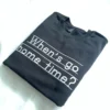 Black crewneck laid at an angle with the white embroidered question When's go home time? on writing guide lines