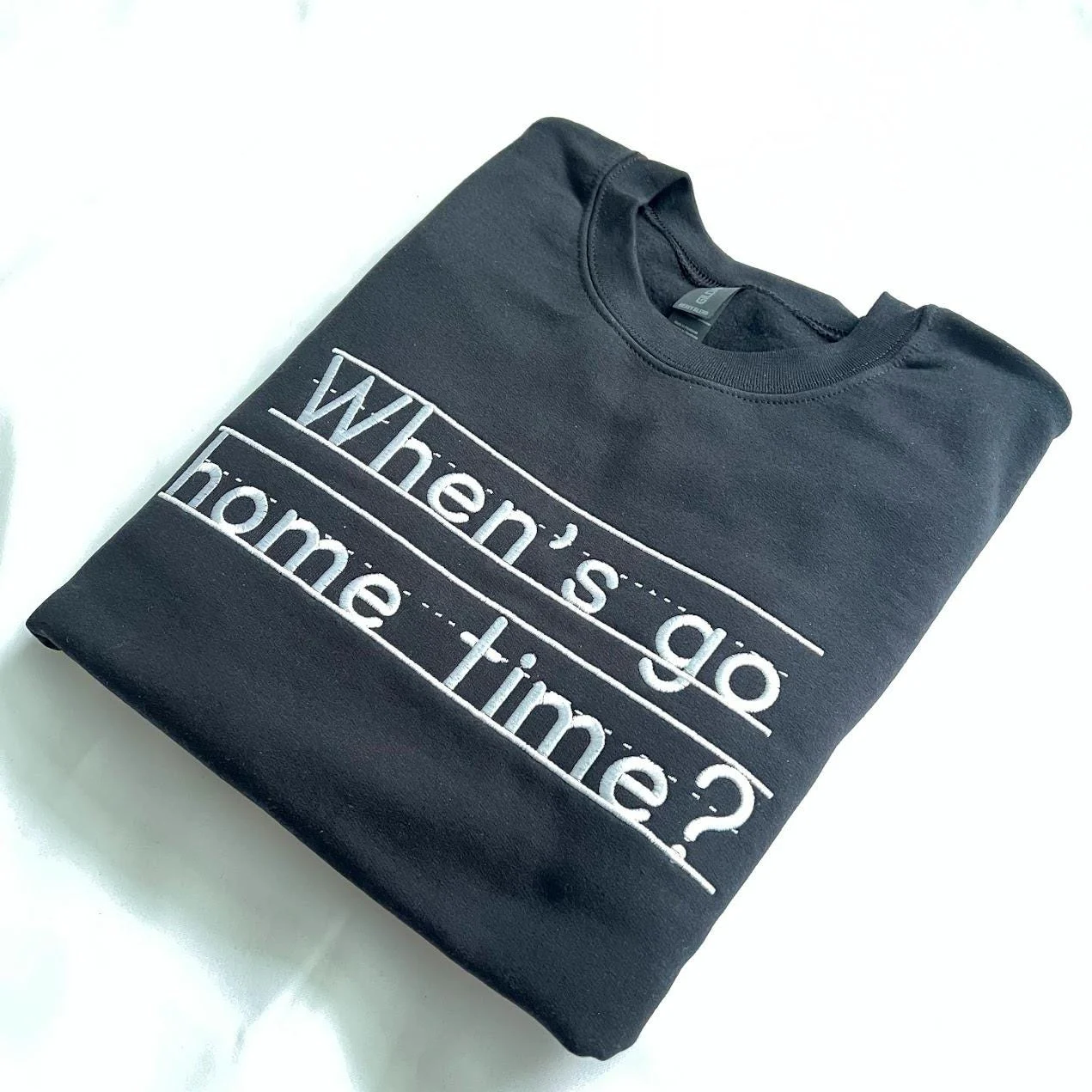 Black crewneck laid at an angle with the white embroidered question When's go home time? on writing guide lines