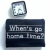 Folded black sweatshirt with When's go home time? embroidered in white print letters on handwriting lines, by a clock