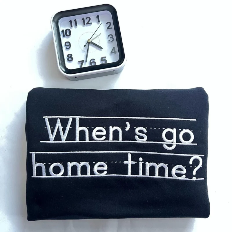 Folded black sweatshirt with When's go home time? embroidered in white print letters on handwriting lines, by a clock