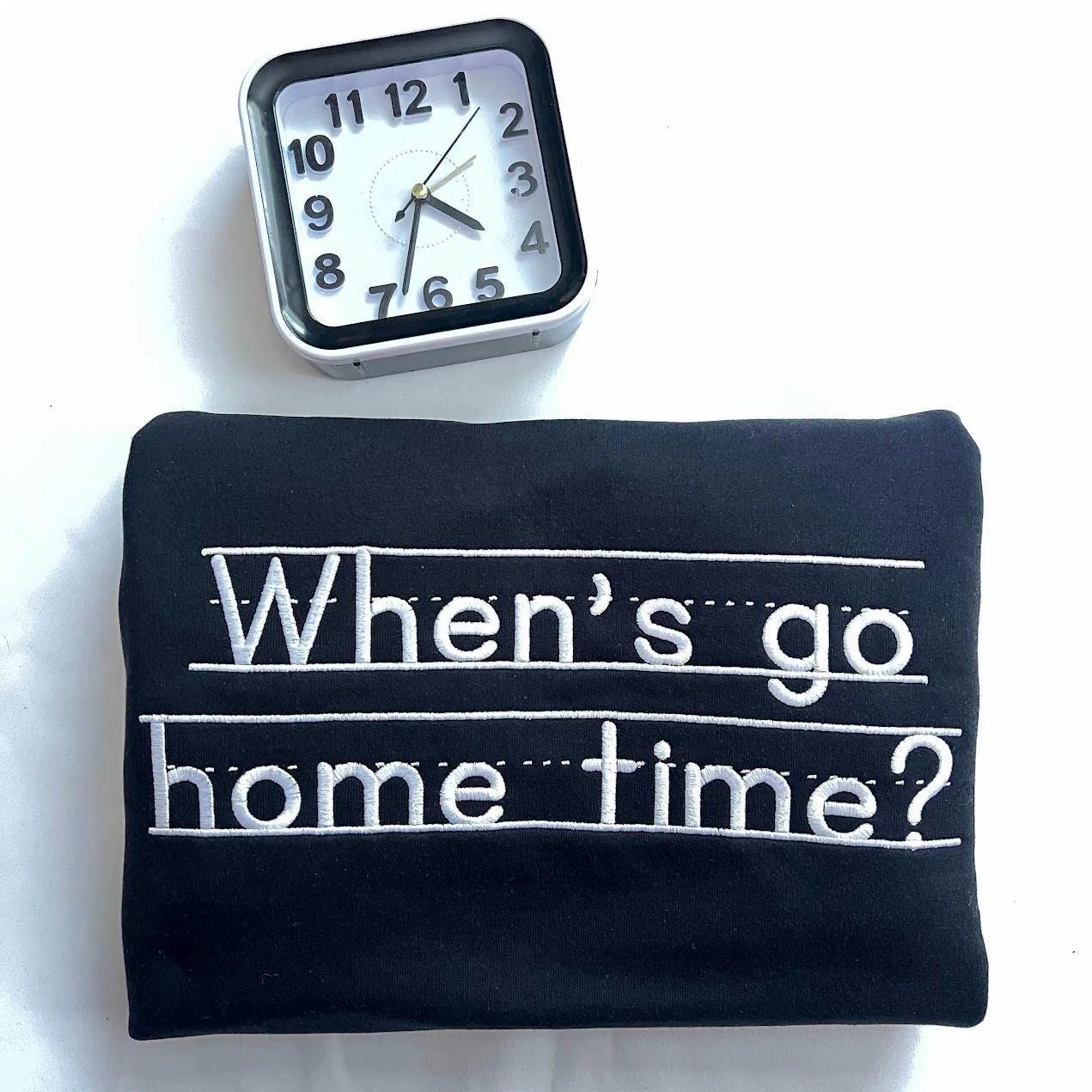 Folded black sweatshirt with When's go home time? embroidered in white print letters on handwriting lines, by a clock