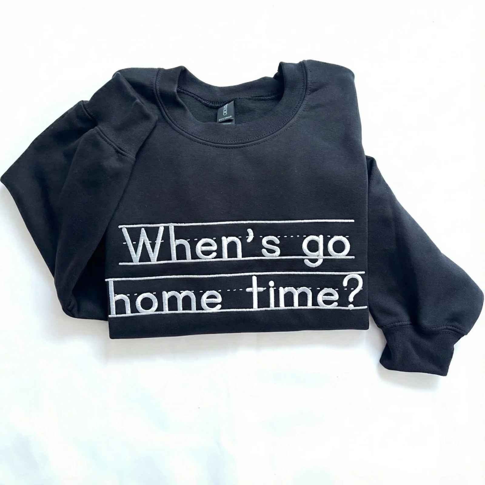 Black sweatshirt flat-lay with When's go home time? stitched in white school-style letters across the chest