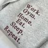 Angled close-up of six stacked maroon serif words ending in Repeat. stitched on an ash grey crewneck