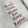 Close-up of the maroon embroidered words Work. Gym. Home. Eat. Sleep. Repeat. on a heather grey sweatshirt