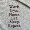 Straight-on detail of the stacked quote Work. Gym. Home. Eat. Sleep. Repeat. in maroon thread on grey