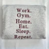 Folded ash grey sweatshirt showing the maroon embroidered quote Work. Gym. Home. Eat. Sleep. Repeat.